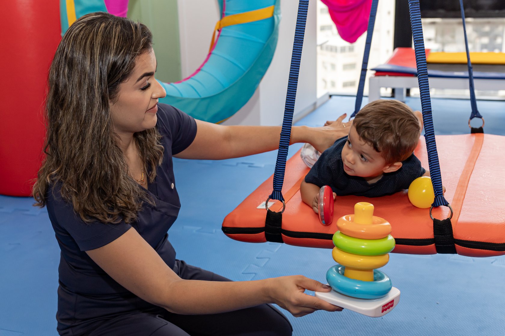 Letícia observando um bebê deitado numa plataforma de balanço enquanto oferece um brinquedo de argolas